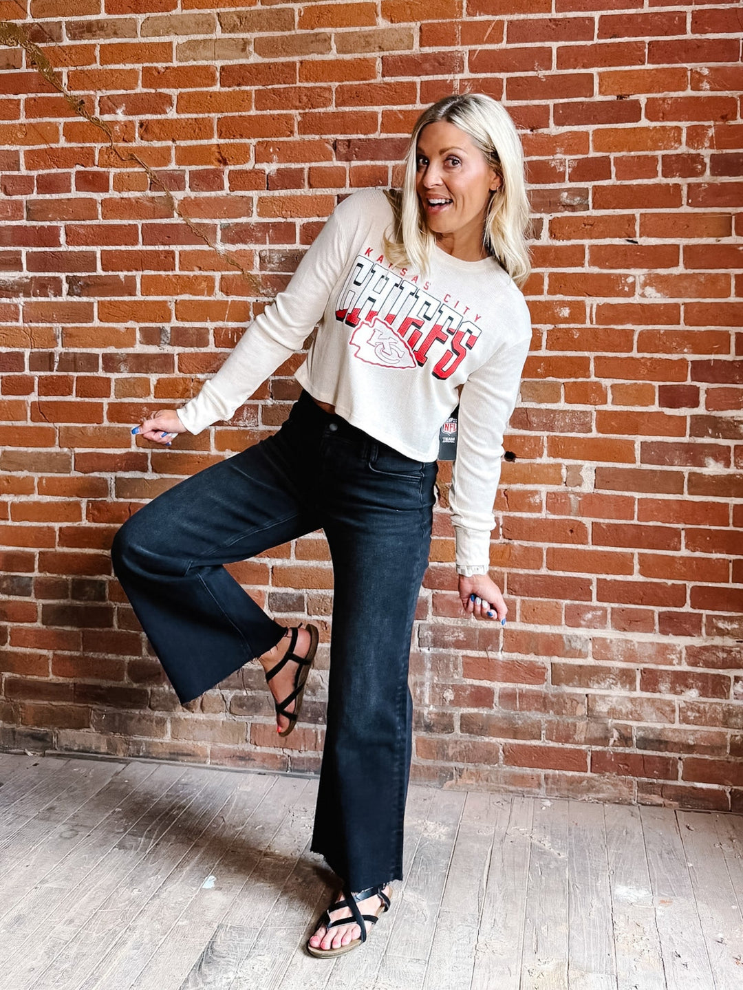 A woman posing in front of a red brick wall wearing a cream long-sleeve graphic sweatshirt, dark blue flare jeans, and black strappy sandals.