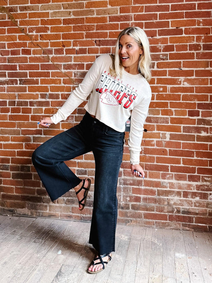 A woman posing in front of a red brick wall wearing a cream long-sleeve graphic sweatshirt, dark blue flare jeans, and black strappy sandals.