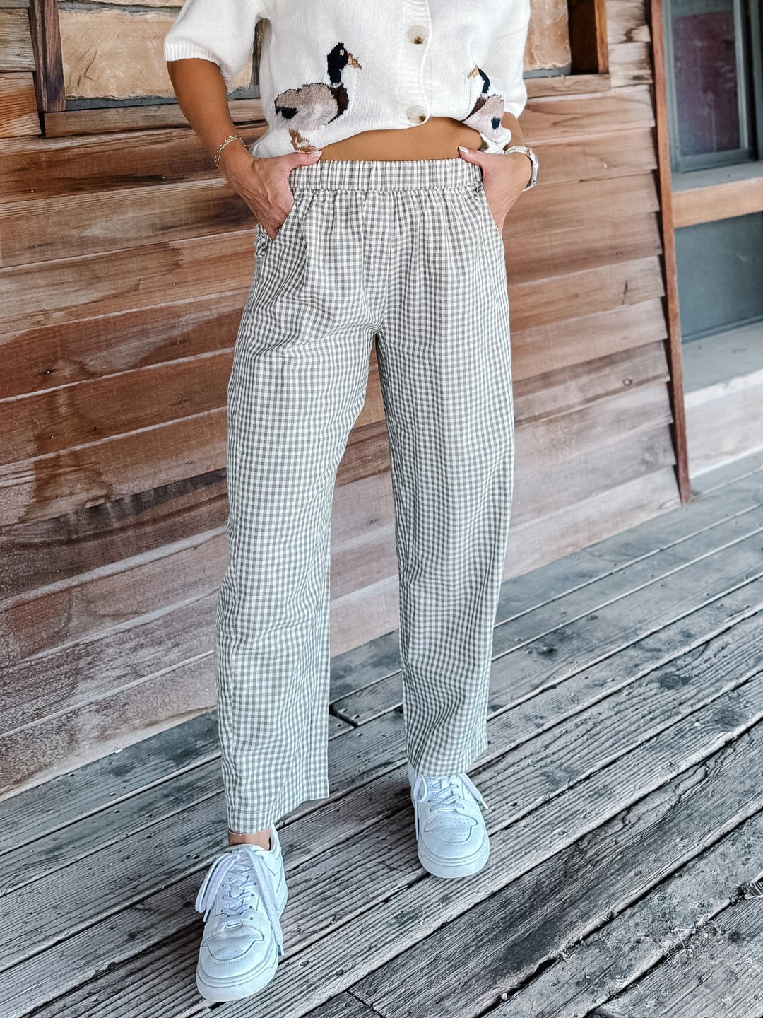 A person wearing loose, light-colored gingham pants with a white cropped button-up shirt and white sneakers, standing on a weathered wooden deck in front of a wood-paneled wall.