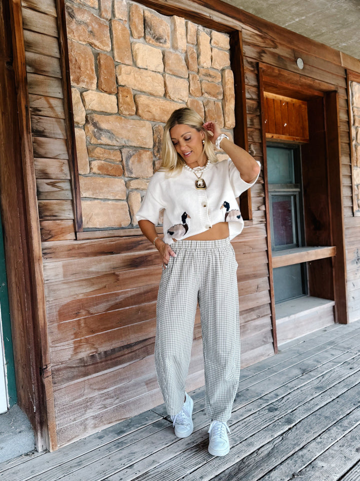 A woman stands on a rustic wooden porch wearing a white cropped short-sleeve top with black and white bird graphics, paired with light gray and white gingham wide-leg pants and white sneakers.
