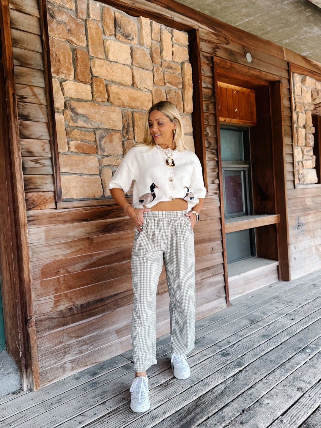 A woman stands on a rustic wooden porch wearing a cream cropped knit top with black and white bird embroidery, light gray and white gingham wide-leg pants, and white sneakers.