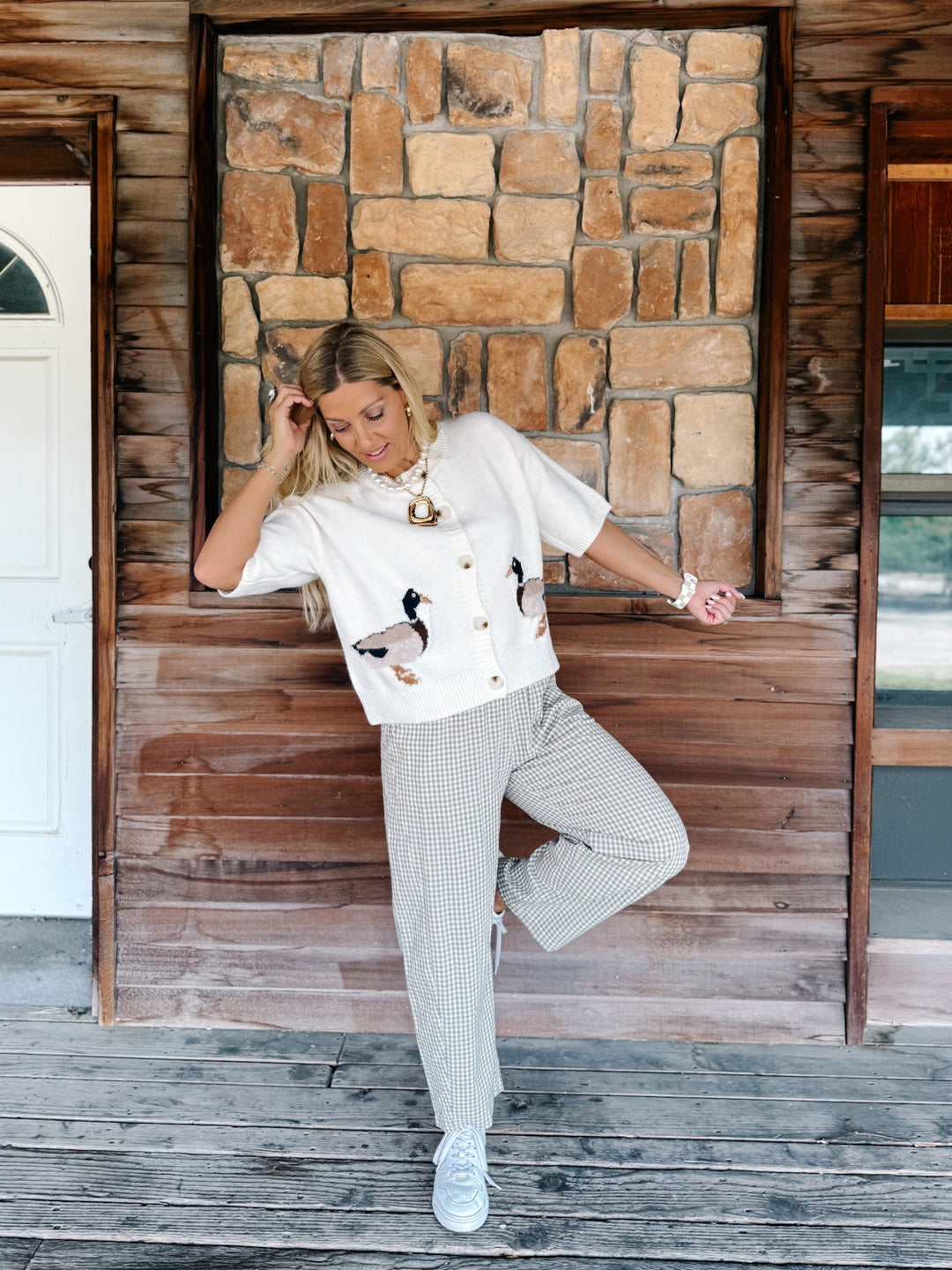 A woman posing on a rustic wooden porch in front of a stone wall, wearing a white short-sleeve button-up shirt with duck embroidery, white and gray patterned trousers, and white sneakers.