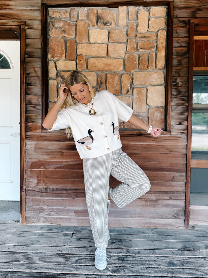 A woman posing on a rustic wooden porch in front of a stone wall, wearing a white short-sleeve button-up shirt with duck embroidery, white and gray patterned trousers, and white sneakers.