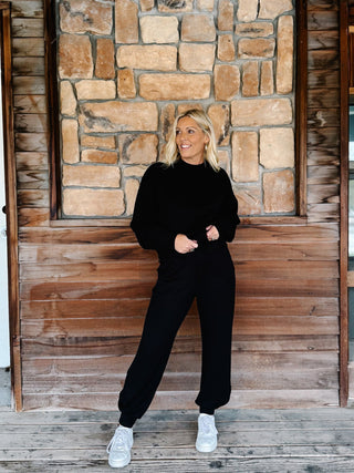 A woman modeling a black two-piece lounge set consisting of a long-sleeve sweatshirt and matching sweatpants, paired with white sneakers, standing on a rustic wooden porch in front of a stone wall.