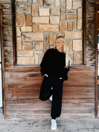 A woman modeling a black two-piece lounge set consisting of a long-sleeve sweatshirt and matching sweatpants, paired with white sneakers, standing on a rustic wooden porch in front of a stone wall.