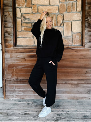 A woman modeling a black two-piece lounge set consisting of a long-sleeve sweatshirt and matching sweatpants, paired with white sneakers, standing on a rustic wooden porch in front of a stone wall.