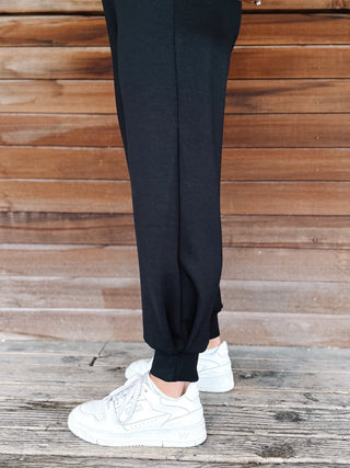 A woman modeling a black two-piece lounge set consisting of a long-sleeve sweatshirt and matching sweatpants, paired with white sneakers, standing on a rustic wooden porch in front of a stone wall.