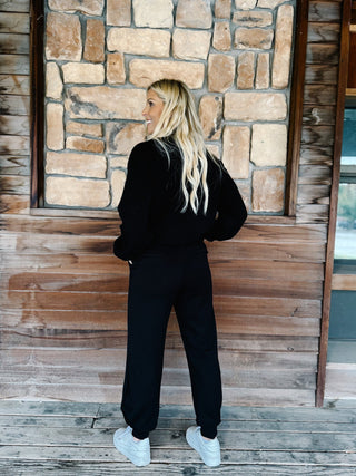 A woman modeling a black two-piece lounge set consisting of a long-sleeve sweatshirt and matching sweatpants, paired with white sneakers, standing on a rustic wooden porch in front of a stone wall.