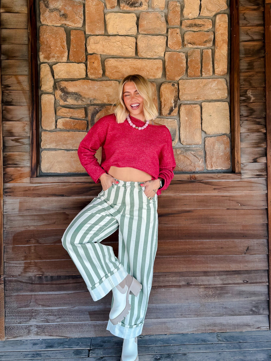 A woman posing in front of a rustic stone and wood wall, wearing a bright pink cropped knit sweater, green and white striped wide-leg pants, and white boots.