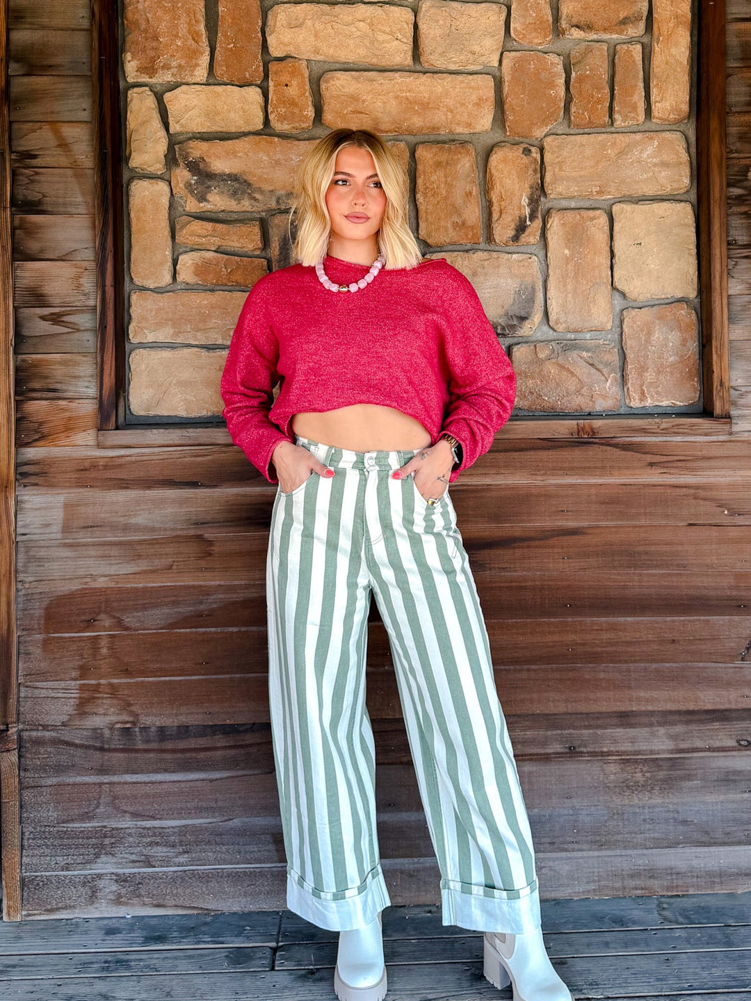 A woman stands in front of a rustic stone and wood wall, wearing a bright pink cropped long-sleeve sweater, light green and white vertically striped wide-leg pants, and white heeled boots.