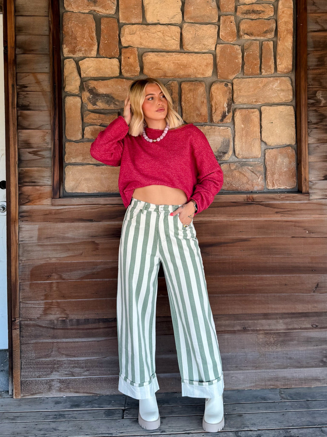 A woman stands on a wooden porch in front of a stone wall, wearing a bright pink cropped long-sleeve sweater, sage green and white vertically striped wide-leg jeans, and white boots.