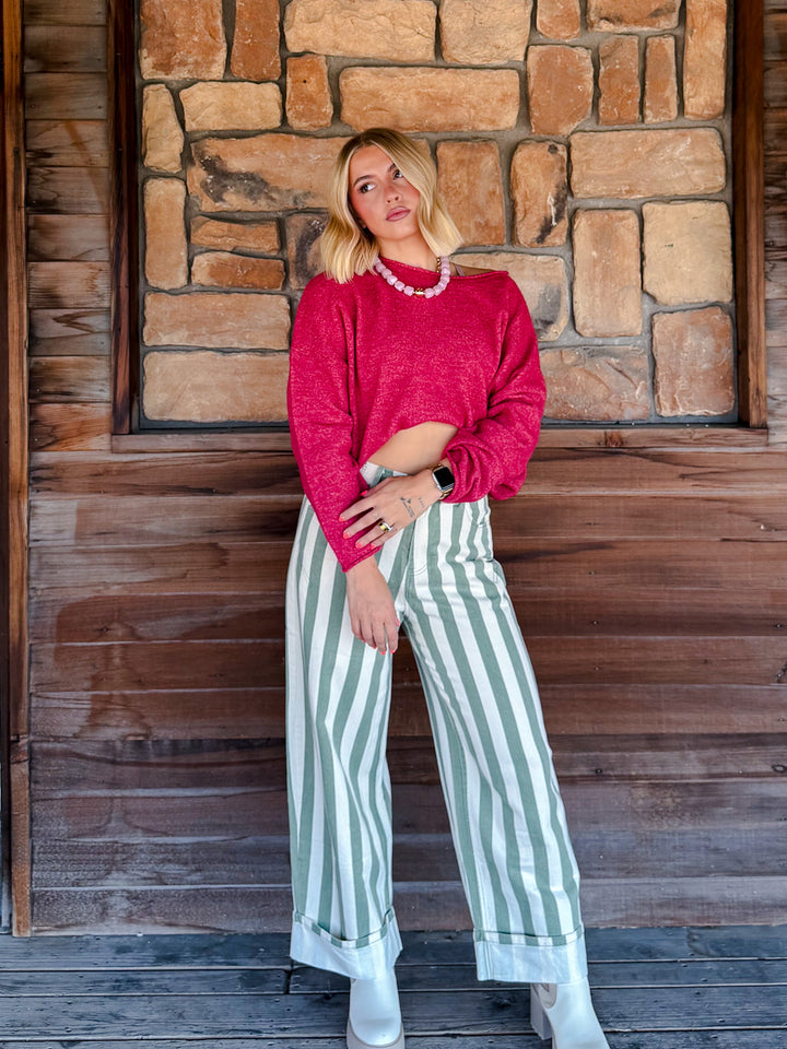 A woman stands in front of a rustic stone and wood wall, wearing a bright pinkish-red knit sweater, green and white vertically striped wide-leg pants, and white boots.
