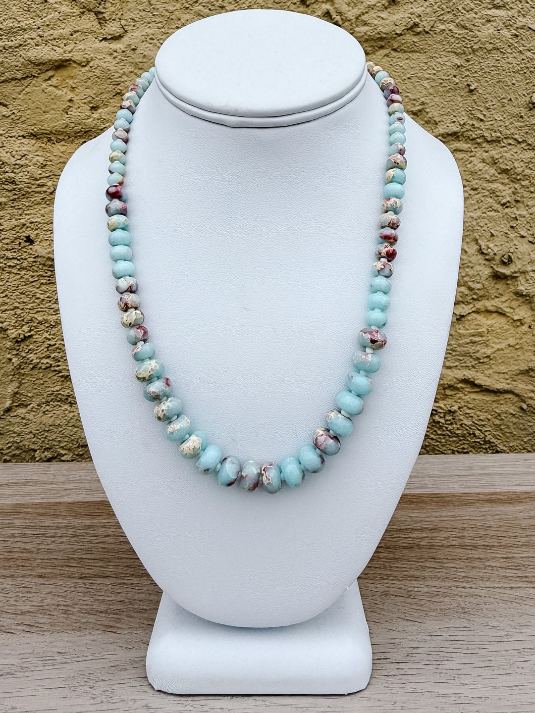 A beaded necklace displayed on a white jewelry bust, featuring light blue turquoise-like beads with small marbled brown and white accents, set against a textured tan wall and light wood surface.