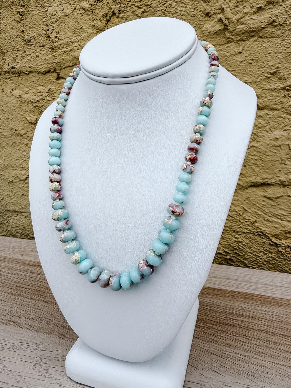 A beaded necklace with light blue turquoise-like beads and marbled red, brown, and white accents, displayed on a white jewelry bust against a textured tan wall.