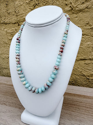 A beaded necklace with light blue turquoise-like beads and marbled red, brown, and white accents, displayed on a white jewelry bust against a textured tan wall.
