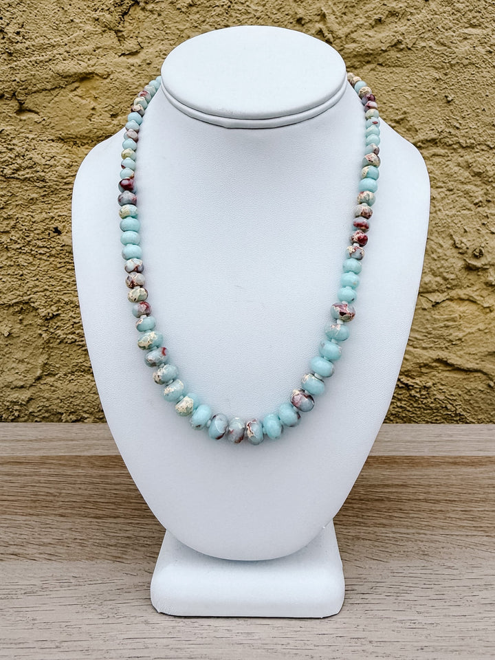 A beaded necklace made of light blue turquoise-like beads with small marbled reddish and cream accents, displayed on a white jewelry bust against a textured tan wall and wood surface.