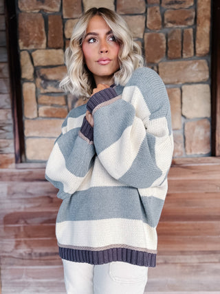A woman wearing a chunky knit sweater with wide horizontal stripes in light blue-gray and cream, posed in front of a rustic stone wall.