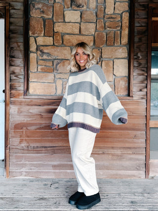 A woman stands on a wooden porch in front of a stone wall, wearing an oversized striped knit sweater with wide sleeves and cream wide-leg pants.