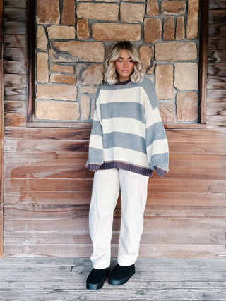 A woman stands on a wooden porch in front of a stone wall, wearing an oversized knit sweater with wide horizontal stripes in light blue-gray and cream, paired with white pants and black shoes.