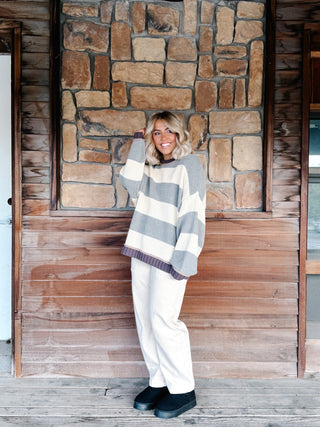 A woman stands on a wooden porch in front of a stone and wood wall, wearing a chunky knit sweater with wide gray and cream horizontal stripes, cream wide-leg pants, and black boots.