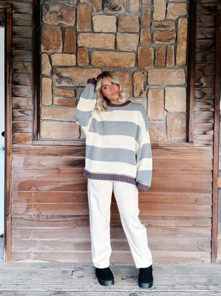 A woman stands on a rustic wooden porch in front of a stone wall, wearing a chunky knit sweater with wide gray and cream horizontal stripes, paired with cream pants and black boots.