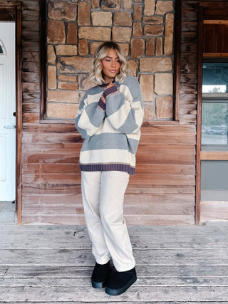A woman stands on a rustic wooden porch in front of a stone and wood wall, wearing a chunky knit sweater with wide gray and cream stripes, cream pants, and black boots.