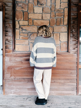A person standing with their back to the camera, wearing a gray and cream striped knit sweater, cream pants, and black shoes, positioned in front of a rustic stone and wood wall.