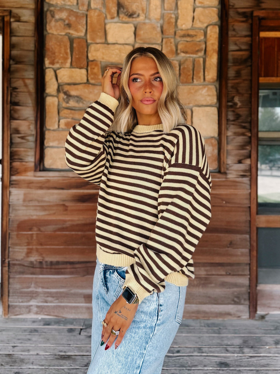 A woman wearing a brown and cream striped knit sweater and light-wash blue jeans, standing on a wooden porch in front of a stone wall.