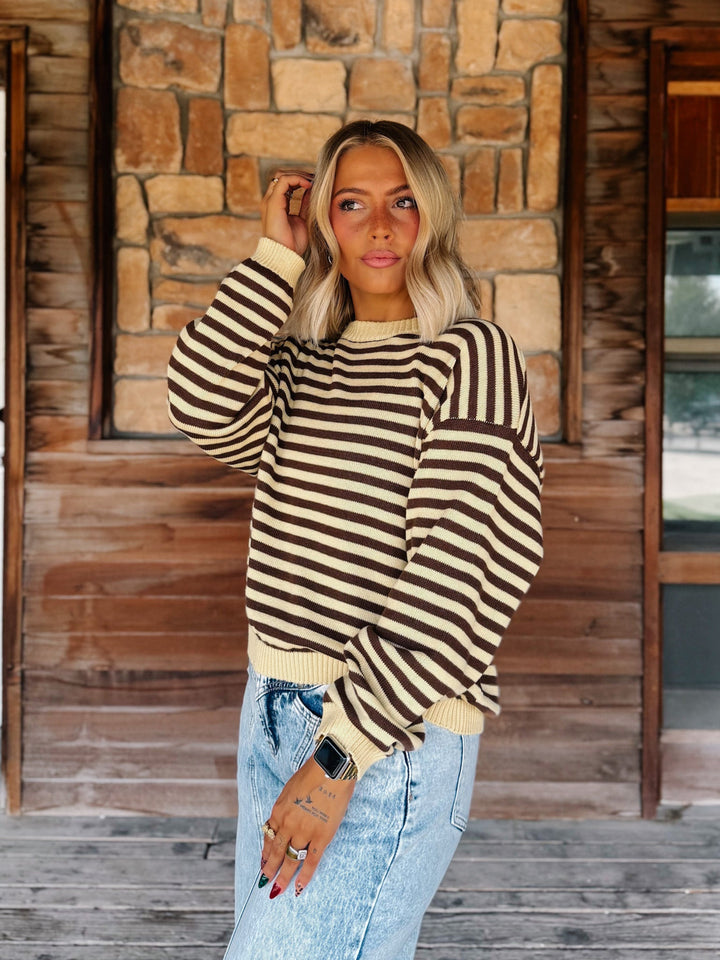A woman wearing a brown and cream striped knit sweater and light-wash blue jeans, standing on a wooden porch in front of a stone wall.