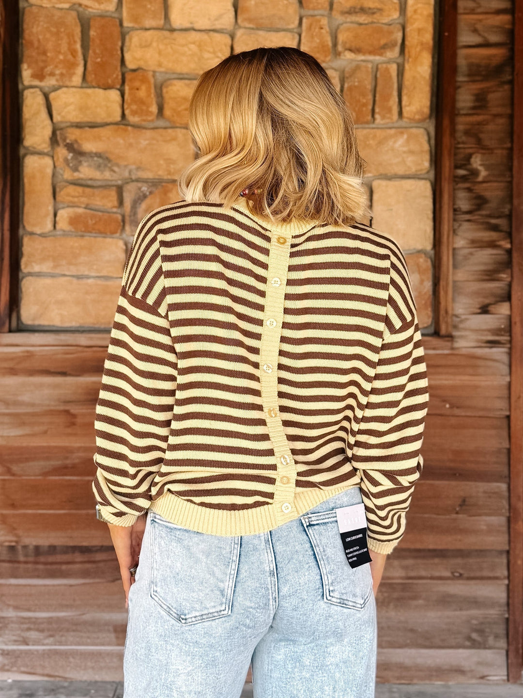 A woman viewed from behind wearing a brown and cream striped button-up cardigan sweater and light wash jeans, standing in front of a rustic stone and wood wall.