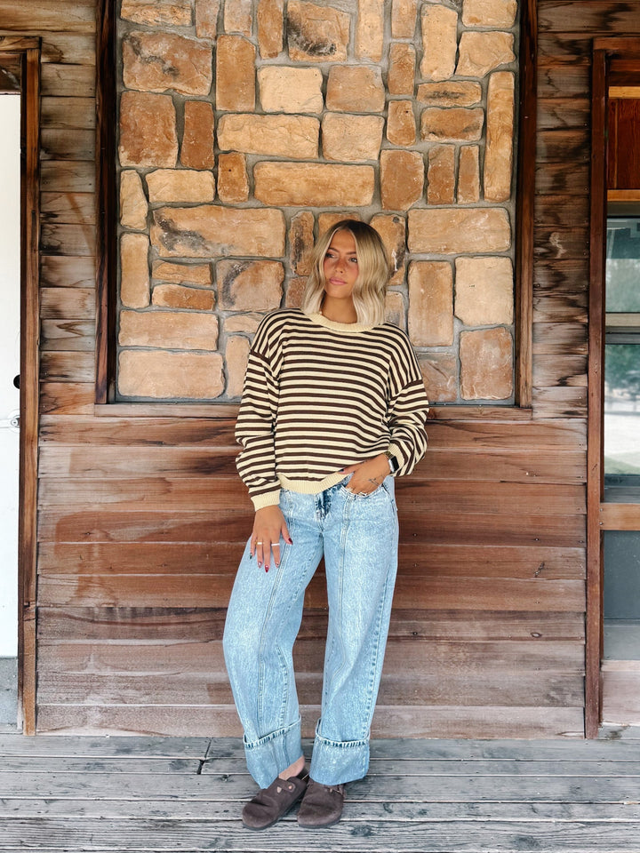 A woman stands on a wooden porch in front of a stone and wood wall, wearing a beige and black striped long-sleeve knit top, light-wash wide-leg jeans, and brown shoes.