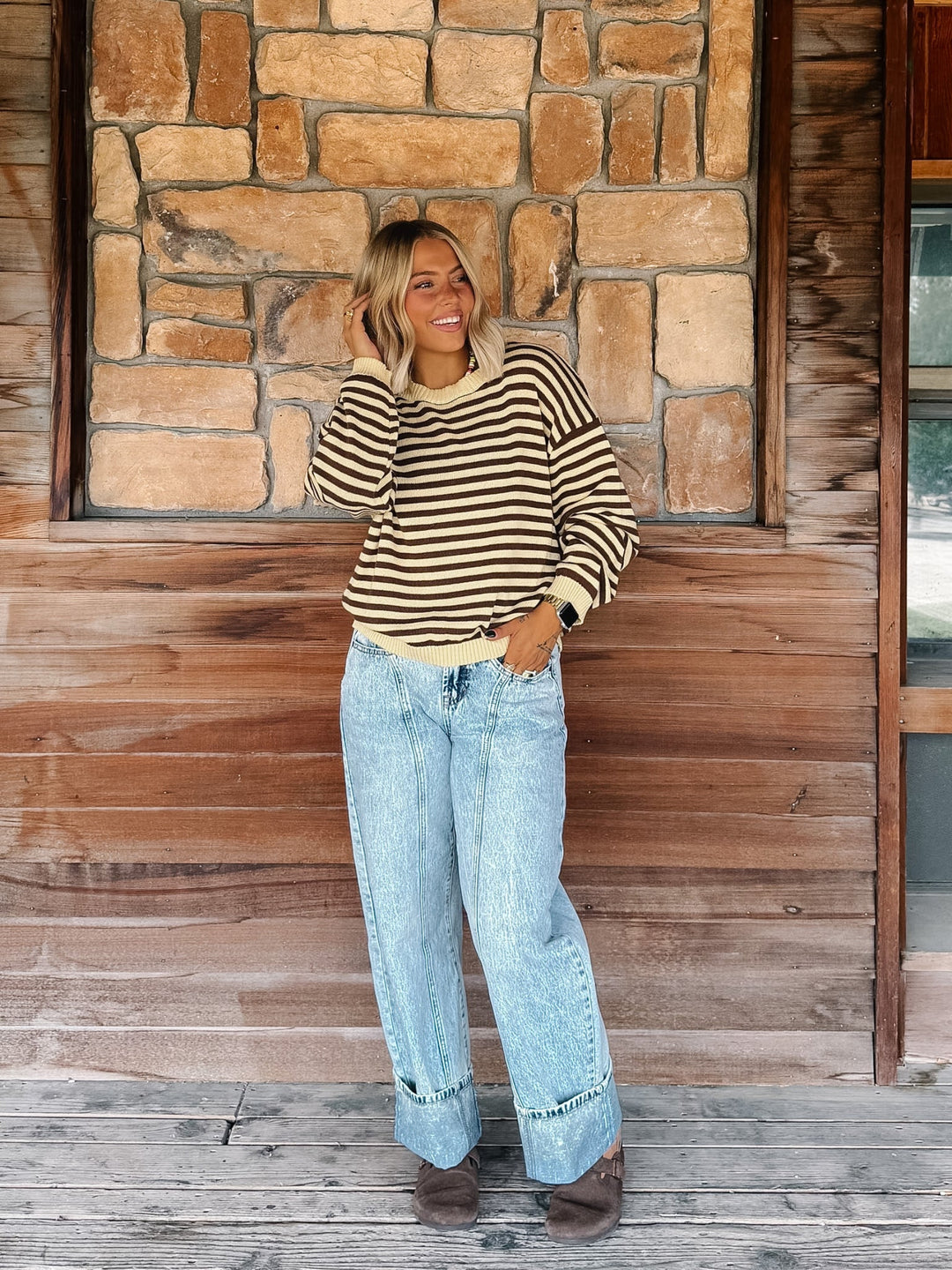 A woman stands on a wooden porch in front of a stone and wood wall, wearing a beige and black striped long-sleeve top, light-wash wide-leg jeans, and brown boots.