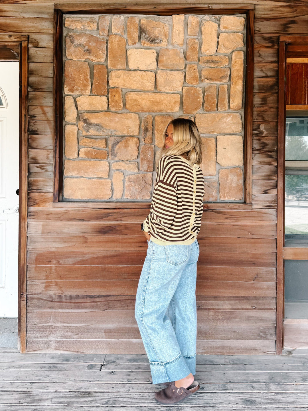 A woman stands on a rustic wooden porch in front of a stone fireplace, wearing a beige and black striped long-sleeve top, light blue wide-leg jeans, and brown slip-on shoes.