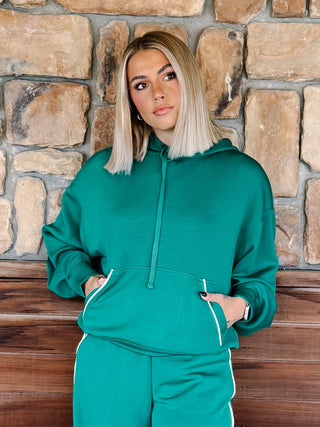 A woman with blonde hair wearing a kelly green hoodie and matching sweatpants with white side stripes, standing in front of a rustic stone wall.