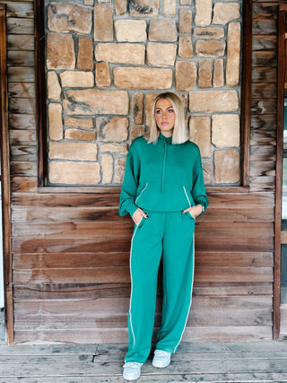 A woman with blonde hair wearing a kelly green hoodie and matching sweatpants with white side stripes, standing in front of a rustic stone wall.