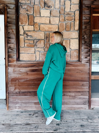 A woman with blonde hair wearing a kelly green hoodie and matching sweatpants with white side stripes, standing in front of a rustic stone wall.