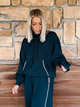 A woman wearing a navy teal matching lounge set consisting of a pullover hoodie and sweatpants with white piping details, standing in front of a rustic stone wall and wooden floor.