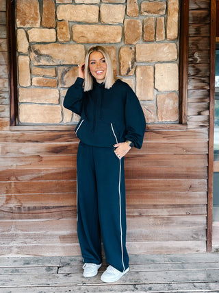 A woman wearing a navy teal matching lounge set consisting of a pullover hoodie and sweatpants with white piping details, standing in front of a rustic stone wall and wooden floor.
