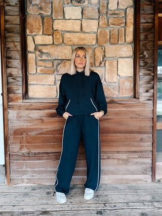 A woman wearing a navy teal matching lounge set consisting of a pullover hoodie and sweatpants with white piping details, standing in front of a rustic stone wall and wooden floor.