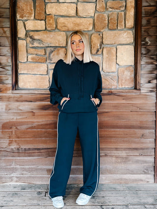 A woman wearing a navy teal matching lounge set consisting of a pullover hoodie and sweatpants with white piping details, standing in front of a rustic stone wall and wooden floor.