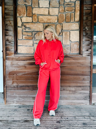 A woman wearing a red two-piece tracksuit with a pullover hoodie and matching sweatpants with white side stripes, standing in front of a stone wall.