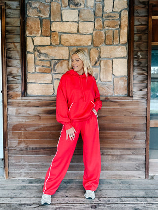 A woman wearing a red two-piece tracksuit with a pullover hoodie and matching sweatpants with white side stripes, standing in front of a stone wall.