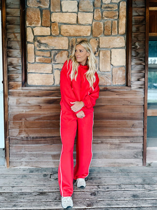 A woman wearing a red two-piece tracksuit with a pullover hoodie and matching sweatpants with white side stripes, standing in front of a stone wall.