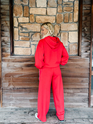 A woman wearing a red two-piece tracksuit with a pullover hoodie and matching sweatpants with white side stripes, standing in front of a stone wall.