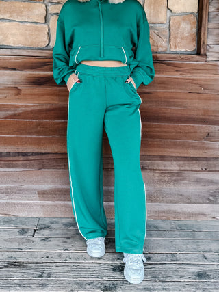 A person wearing a matching green hoodie and wide-leg sweatpants with white side stripes, standing against a rustic wood and stone wall.