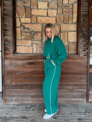 A woman wearing a matching green  hoodie and wide-leg sweatpants with white side stripes, standing on a wooden deck in front of a stone wall.