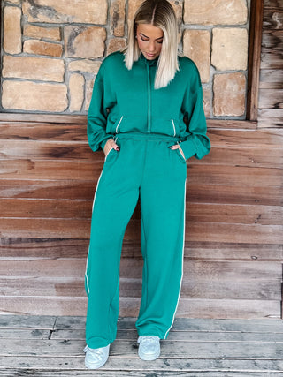 A woman wearing a matching green zip-up hoodie and wide-leg sweatpants with white side stripes, standing against a rustic wooden wall and stone background.