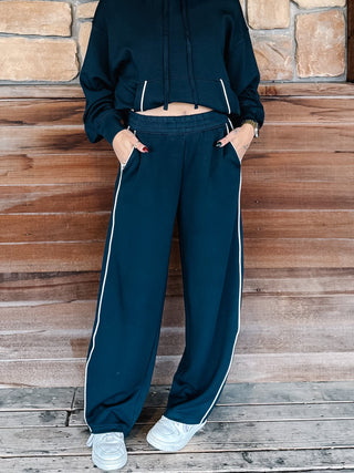 A person wearing teal navy wide-leg sweatpants with white side stripes, paired with a matching hoodie and white sneakers, standing against a rustic wooden wall.