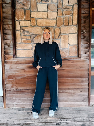 A person wearing teal navy wide-leg sweatpants with white side stripes, paired with a matching hoodie and white sneakers, standing against a rustic wooden wall.