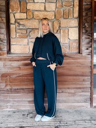 A person wearing teal navy wide-leg sweatpants with white side stripes, paired with a matching hoodie and white sneakers, standing against a rustic wooden wall.